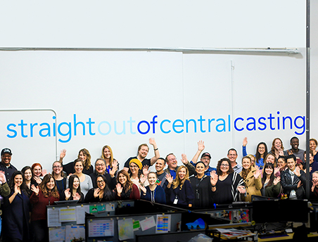 Central Casting staff members waving in front of straight out of Central Casting sign.